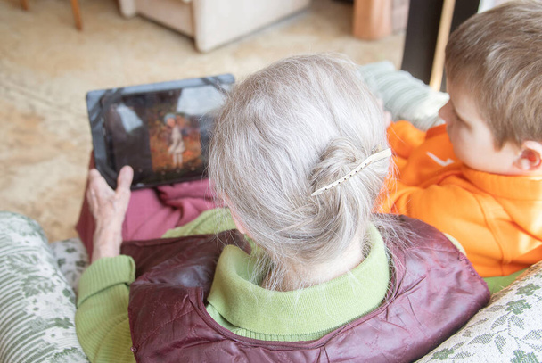 Une vieille femme européenne avec un garçon mignon dans un raglan orange est assis sur une chaise avec une tablette et bavarder en ligne. - Photo, image