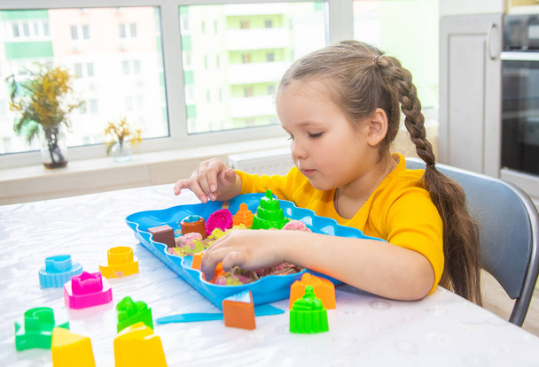 Une petite fille européenne joue avec des perles dans le sable coloré, l'enfant est engagé dans le jeu créatif, le développement précoce, la motricité fine, les jeux pour enfants à la maison. - Photo, image