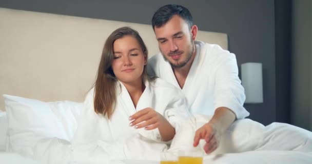 Paar in badjassen drinken sinaasappelsap liggend op groot bed - Video