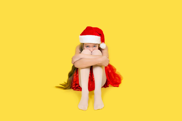Сумна дівчина-підліток у червоній сукні і червоний капелюх Санта-Клауса сидять і обіймають її коліна. Дівчата в Санта - Каті ізольовані на жовтому тлі.. - Фото, зображення