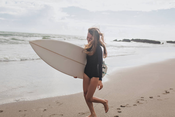 Pretty lovely girl in wetsuit with surfboard walking down the beach in sunlight. High quality photo - Photo, Image