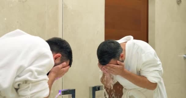 Hombre con barba en albornoz blanco lava la cara en el baño - Imágenes, Vídeo