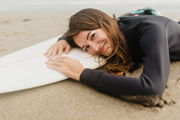 Close up retrato de encantadora mulher de fato de mergulho deitado na prancha de surf na praia de areia em dia quente ensolarado. Foto de alta qualidade - Foto, Imagem