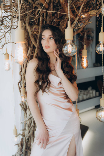Beautiful, stylish, and elegant girl brunette, model, with makeup and curly hair in a peach-colored silk dress for a magazine photoshoot in fashion style. - Valokuva, kuva