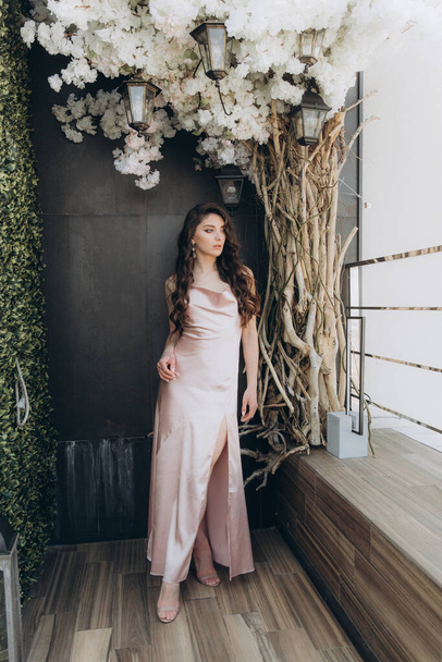 Beautiful, stylish, and elegant girl brunette, model, with makeup and curly hair in a peach-colored silk dress for a magazine photoshoot in fashion style. - Φωτογραφία, εικόνα