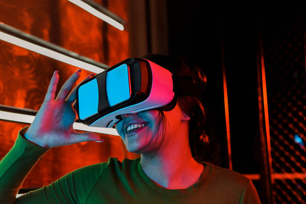 Mujer feliz con auriculares VR sonriendo y explorando la realidad virtual bajo la iluminación de neón rojo en una habitación futurista - Foto, Imagen