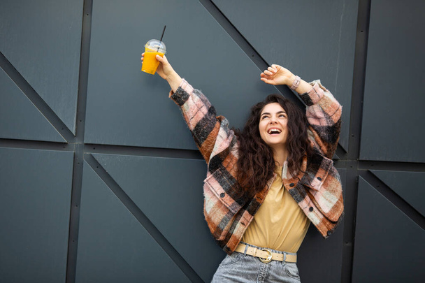 Young stylish woman with juice cup standing over gray background. Portrait of a smiling woman with curly hair. - Φωτογραφία, εικόνα