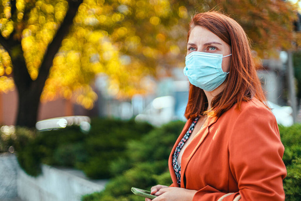 Şehirde duran güvenlik maskeli bir iş kadını. Güzel bir genç kız, kızıl saçlı, Allegria ve virüse karşı koruyucu bir tıbbi maske takıyor. - Fotoğraf, Görsel