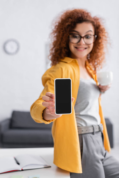 alegre freelancer mostrando teléfono móvil con pantalla en blanco, fondo borroso - Foto, Imagen