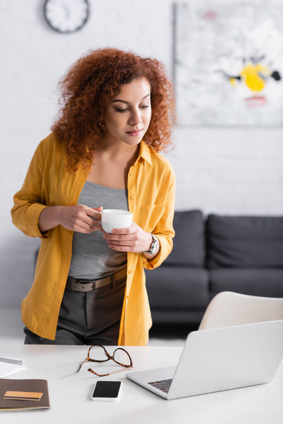 jovem freelancer segurando xícara de café enquanto olha para laptop na mesa - Foto, Imagem