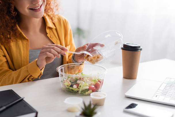 partial view of smiling teleworker holding plastic fork near fresh salad and takeaway drink - Foto, Imagem