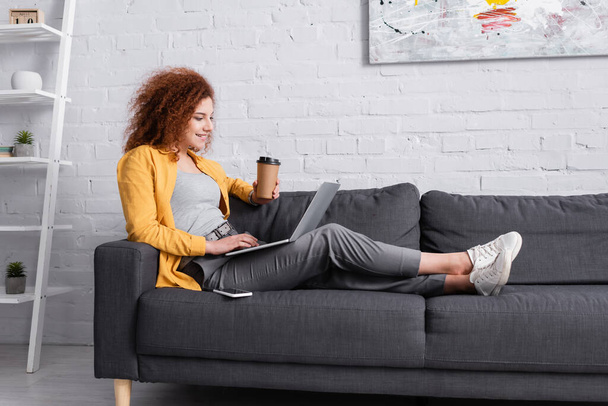 smiling, curly woman sitting on couch with laptop and coffee to go  - Foto, immagini
