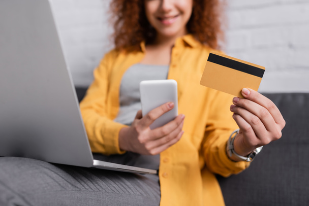 partial view of woman holding smartphone and credit card near laptop on blurred background - Zdjęcie, obraz