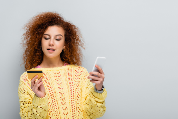 curly woman in knitted sweater holding smartphone and credit card isolated on grey - Fotó, kép