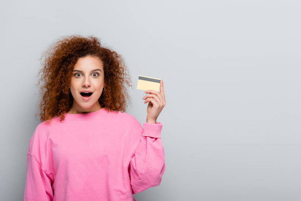 surprised woman looking at camera while holding credit card isolated on grey - Фото, изображение