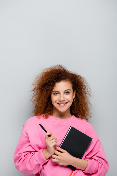 curly woman with notebook and pen smiling at camera isolated on grey - Foto, Bild