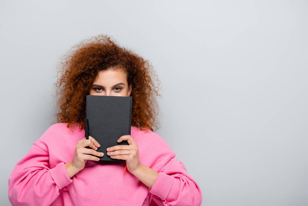curly woman obscuring face with notebook while looking at camera isolated on grey - 写真・画像