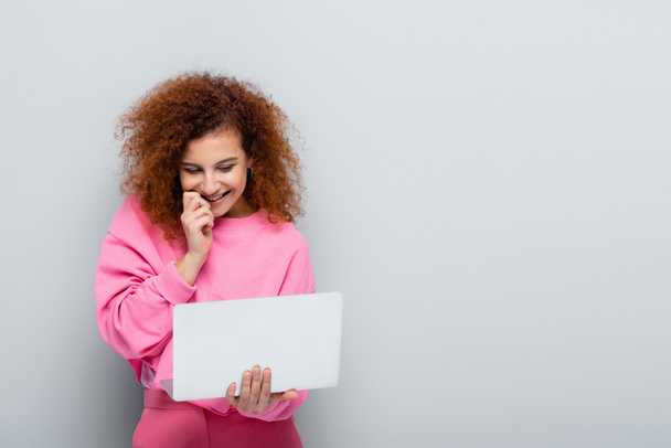 cheerful woman holding hand near face while looking at laptop on grey background - Foto, afbeelding