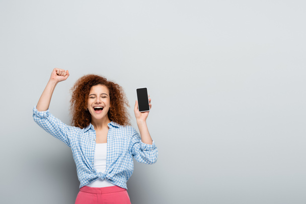 mujer alegre con los ojos cerrados mostrando gesto de regocijo mientras sostiene el teléfono inteligente sobre fondo gris - Foto, imagen