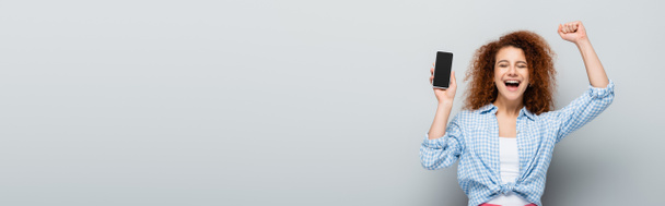 radosna kobieta pokazująca gest sukcesu trzymając telefon komórkowy z pustym ekranem na szarym tle, baner - Zdjęcie, obraz
