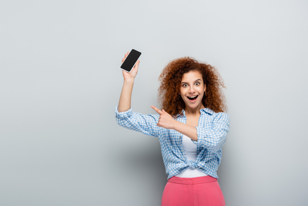 astonished woman pointing with finger while holding smartphone with blank screen on grey background - Photo, image