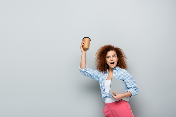 aufgeregte Frau hält Kaffee to go in der Hand, während sie Laptop auf grauem Hintergrund trägt - Foto, Bild