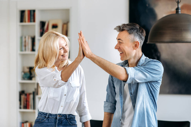 Mature happy caucasian man and woman, business partners, standing in the office, in casual clothes, looking and giving five to each other, gesture of success, smiling happily, deal, agreement - Photo, Image