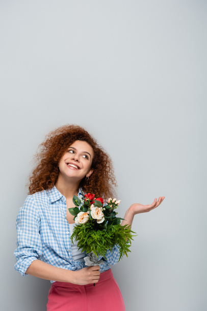 Neşeli kadın, gri arka planda çiçekleri tutarken elini kaldırıp işaret ediyor. - Fotoğraf, Görsel