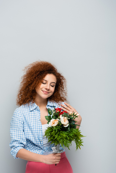 pleased woman with curly hair holding flowers on grey background - Fotoğraf, Görsel