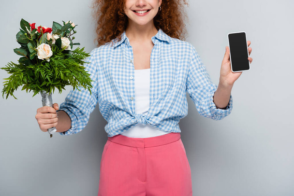 cropped view of smiling woman holding flowers and smartphone with blank screen on grey background - Photo, image