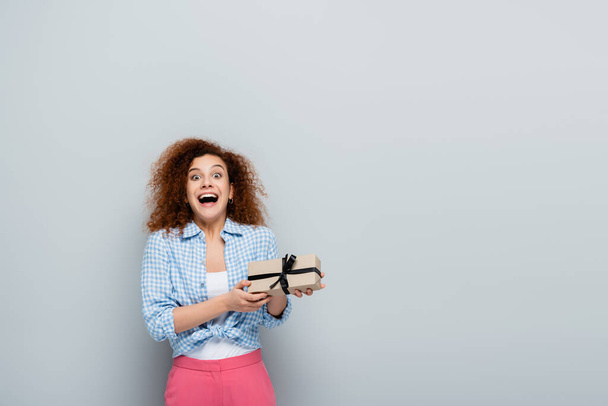 excited woman with open mouth looking at camera while holding present on grey background - Foto, immagini