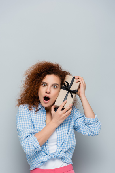 astonished woman with open mouth holding gift box on grey background - Φωτογραφία, εικόνα