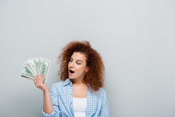 ενθουσιασμένοι γυναίκα σε καρό πουκάμισο κοιτάζοντας χαρτονομίσματα δολάριο απομονώνονται σε γκρι - Φωτογραφία, εικόνα