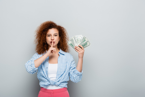 kudrnatá žena ukazující tiché gesto, zatímco drží dolary na šedém pozadí - Fotografie, Obrázek