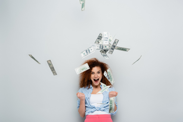 opgewonden vrouw schreeuwen en tonen succes gebaar in de buurt van dollars vallen op grijze achtergrond - Foto, afbeelding