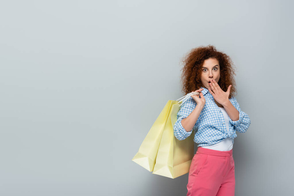изумленная женщина закрывает рот рукой, держа пакеты для покупок на сером фоне - Фото, изображение