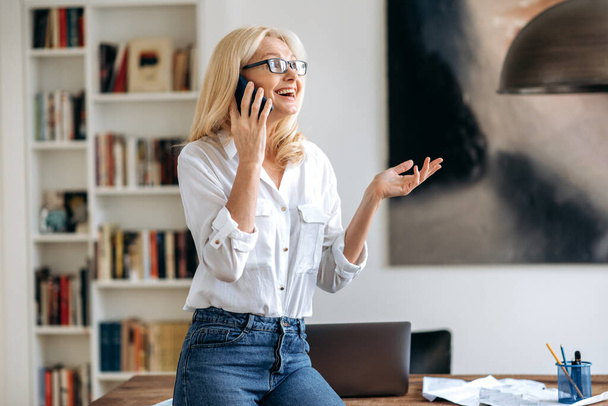Happy friendly busy mature caucasian woman, in stylish wear and glasses, working at office, standing near desk, talking to employee or client on the cell phone, gesturing with hand, smiling - Photo, Image