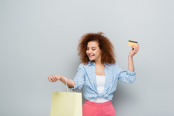 curly woman smiling while holding credit card and shopping bag on grey background - Fotografie, Obrázek