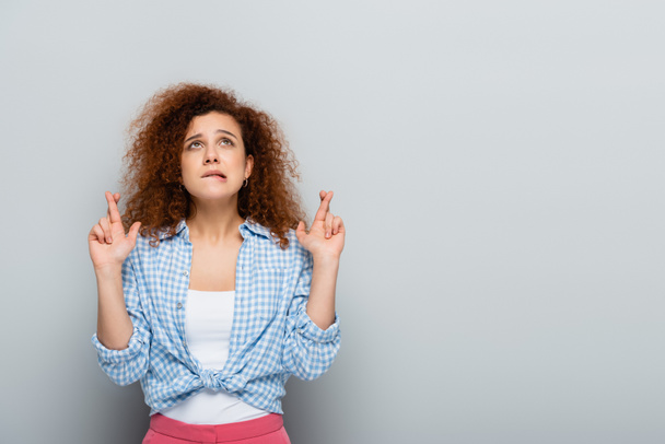 worried woman in plaid shirt looking up while holding crossed fingers on grey background - Foto, Bild