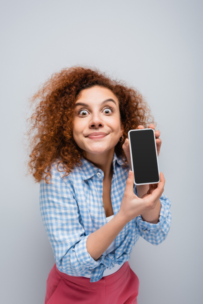 mujer emocionada haciendo muecas mientras muestra teléfono inteligente con pantalla en blanco aislado en gris - Foto, imagen