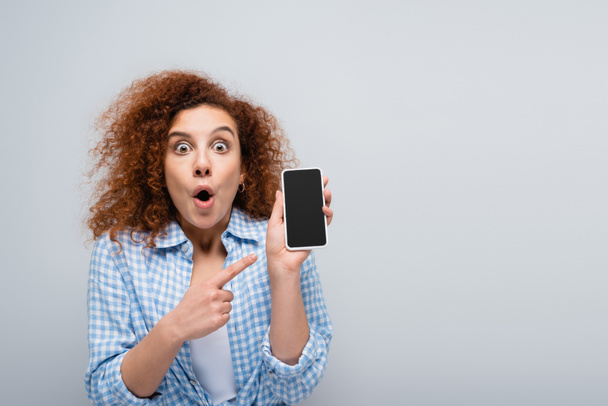 astonished woman looking at camera while pointing at cellphone with blank screen isolated on grey - Foto, Imagem