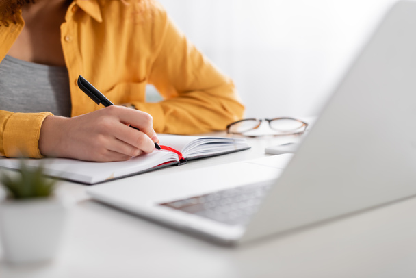 περικοπή άποψη του freelancer γράφοντας στο σημειωματάριο κοντά στο φορητό υπολογιστή σε θολή πρώτο πλάνο - Φωτογραφία, εικόνα