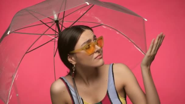 Sad woman in sunglasses holding umbrella isolated on pink - Footage, Video