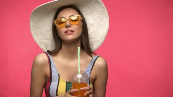 Güneş gözlüklü ve güneş şapkalı kadın içkisini pembe üzerine izole edilmiş halde tutuyor. - Video, Çekim