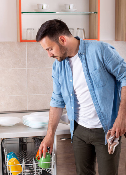 Schöner Mann entlädt Geschirrspüler und wischt Tasse mit Tuch in Küche zu Hause - Foto, Bild