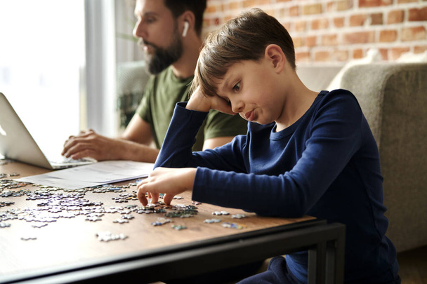 Boredom boy solving jigsaw puzzle during father working                                - Photo, Image