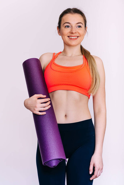 Aantrekkelijke jonge sportvrouw die een fitnessmat vasthoudt terwijl ze geïsoleerd over een witte achtergrond staat. Fitness gezond en sportief concept. - Foto, afbeelding