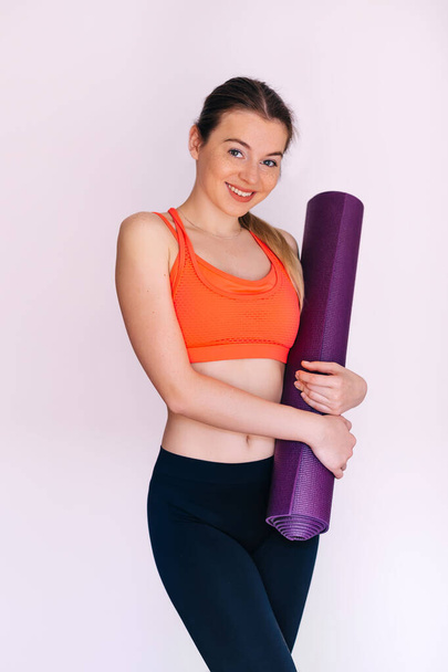 Mooie jonge vrouw in sportkleding houdt violette yoga mat, kijken naar de camera en glimlachen, staan op witte achtergrond - Foto, afbeelding