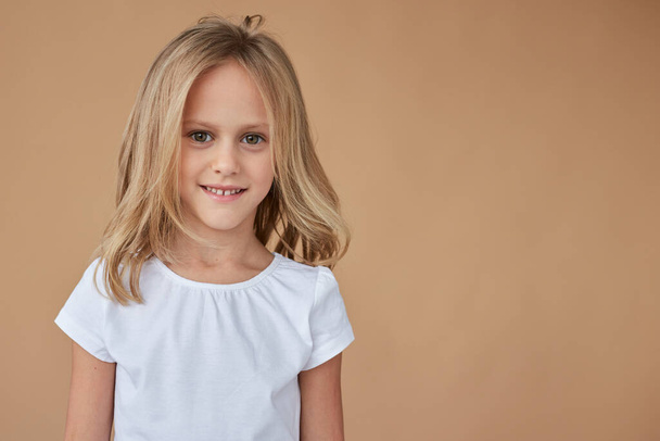 Close-up front portrait of pretty little girl with wavy blond hair, dressed in white clothes, over beige background. Space for text. - Φωτογραφία, εικόνα