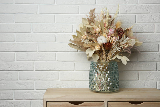 Beautiful dried flower bouquet in glass vase on wooden table near white brick wall. Space for text - Foto, afbeelding